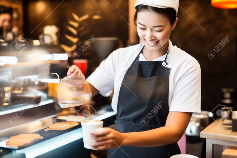 Lächelnde asiatische Barista junge Frau gießt heißen schwarzen Kaffee in Pappbecher für gemäß der Bestellung des Kunden an der Theke im Café.