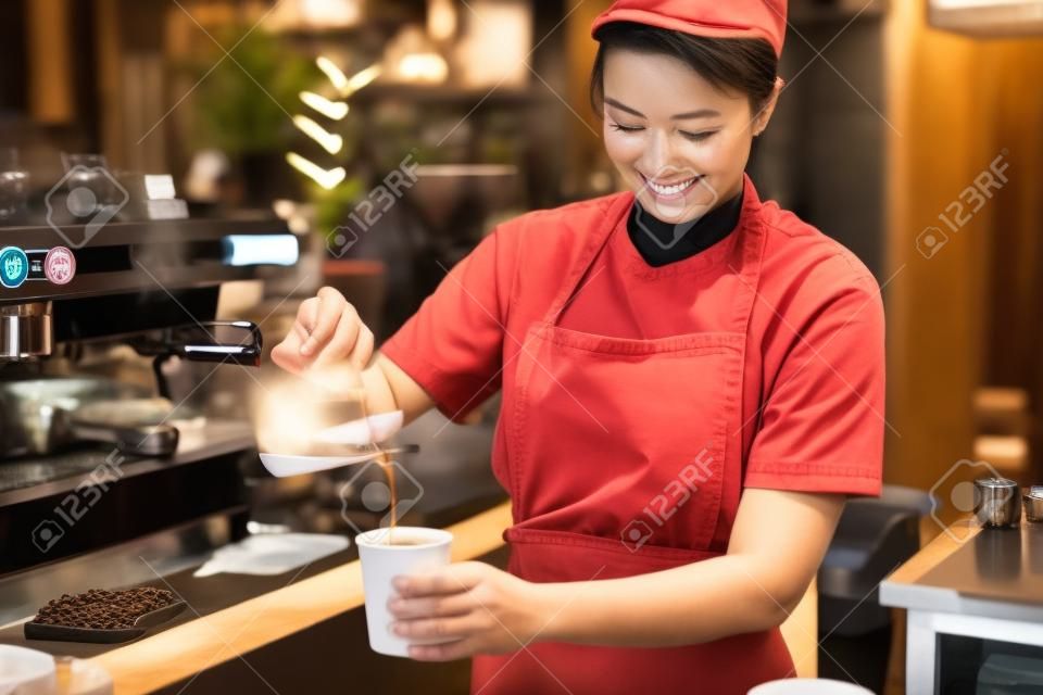 Smiling Asian barista young woman pouring hot black coffee into paper cup for according to the customer's order at counter bar in coffee shop.
