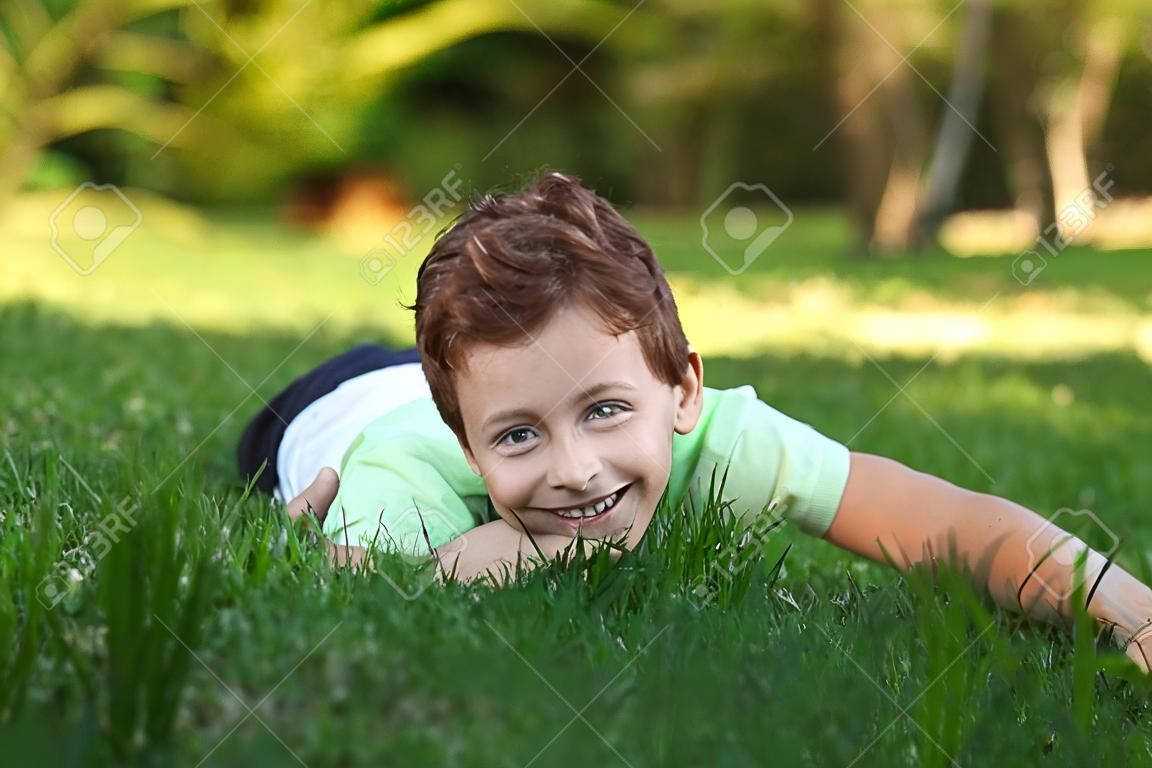 Ein lächelnder kleiner Junge, der auf dem Gras liegt, genießt im Park, schaut an einem sonnigen Sommertag auf die Kamera.