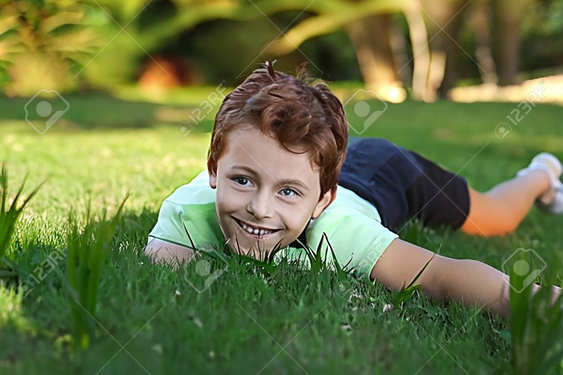 Ein lächelnder kleiner Junge, der auf dem Gras liegt, genießt im Park, schaut an einem sonnigen Sommertag auf die Kamera.