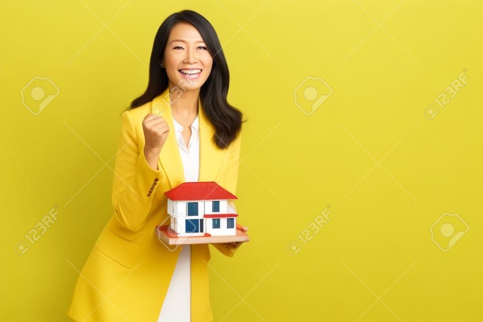 Donna d'affari asiatica sorridente e azienda modello di esempio di casa isolato su sfondo giallo, concetto di assicurazione immobiliare e casa