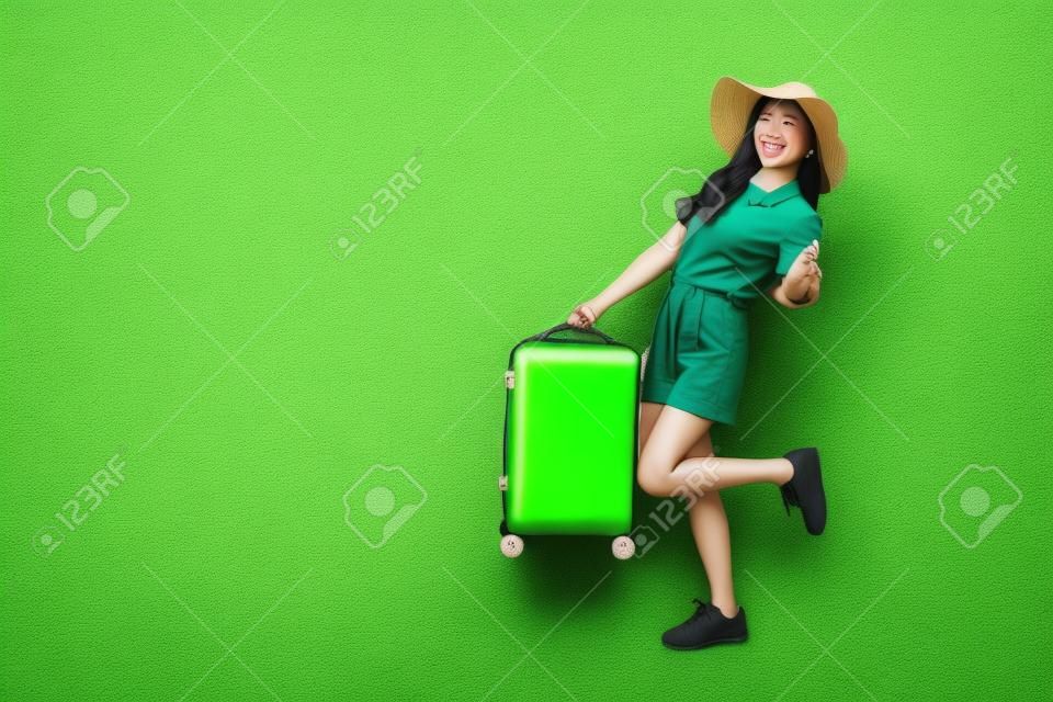 Szczęśliwa azjatycka podróżniczka stojąca i trzymająca walizkę odizolowaną na zielonym tle, turystka mająca wesołą koncepcję podróży wakacyjnej, pełny skład ciała