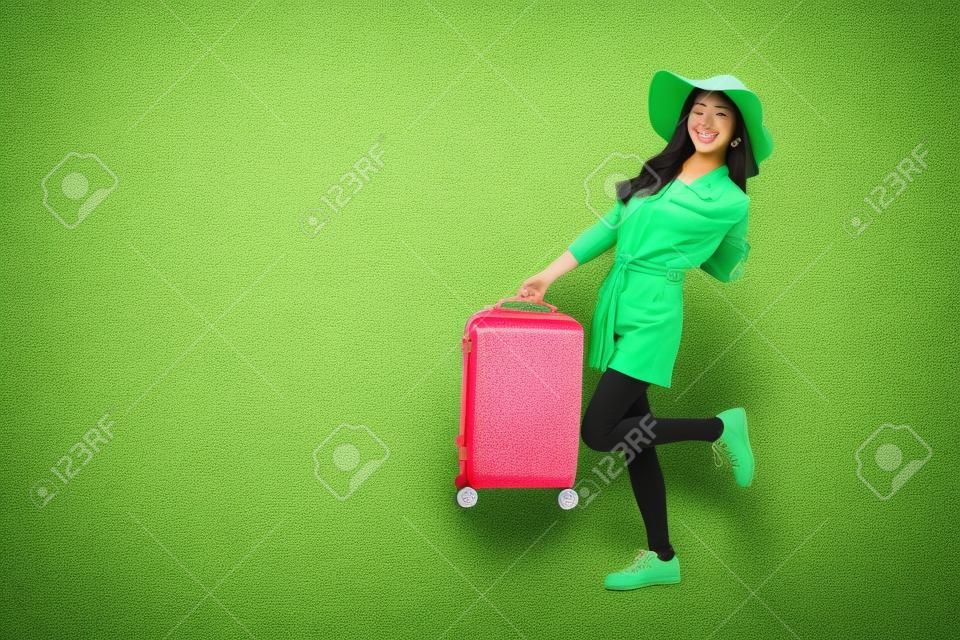 Szczęśliwa azjatycka podróżniczka stojąca i trzymająca walizkę odizolowaną na zielonym tle, turystka mająca wesołą koncepcję podróży wakacyjnej, pełny skład ciała