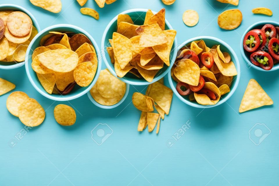 Potato and tortilla chips and other salty snacks, shot from above with copy space. Party food on a teal blue background. A mix of appetizers in bowls