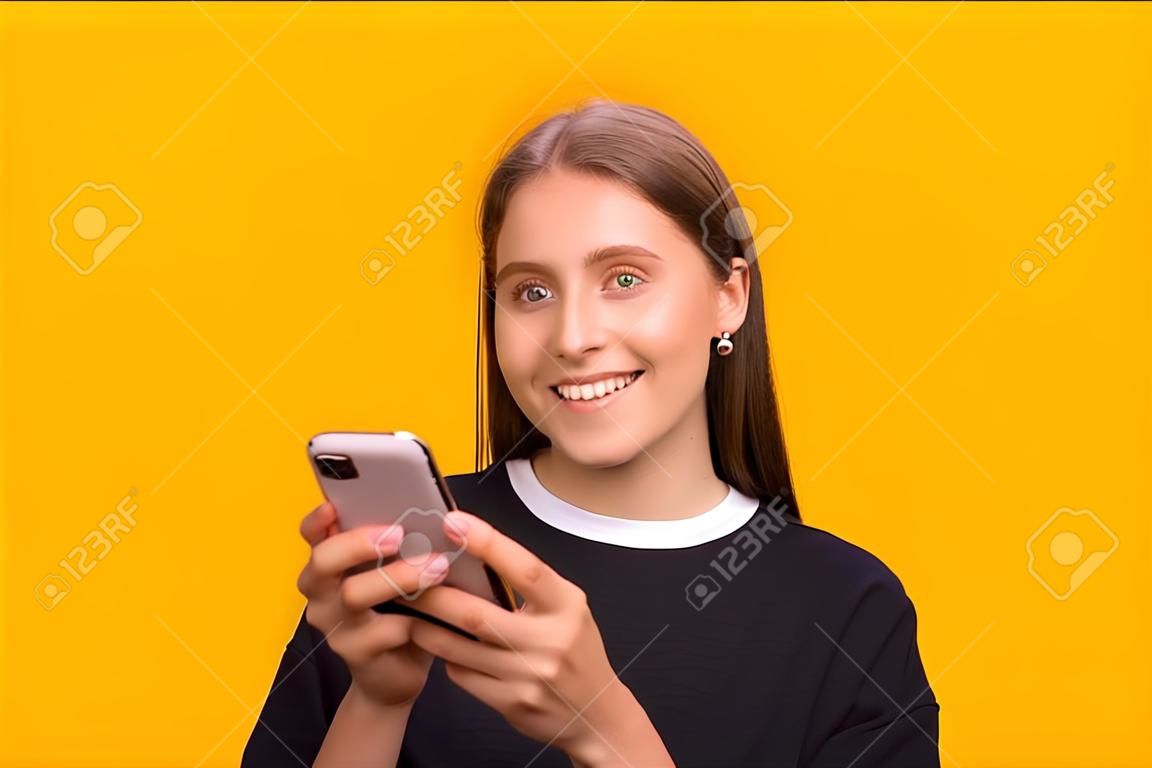 Señora bonita feliz que usa el teléfono móvil, sonriendo a la cámara, de pie contra el fondo amarillo.