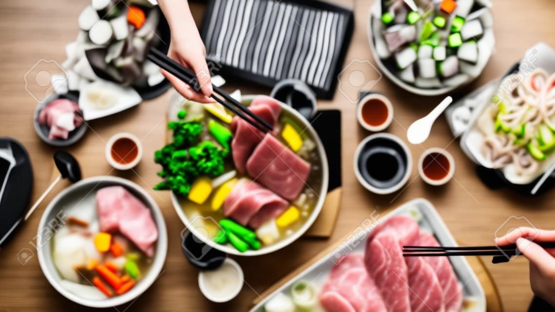신선한 얇게 썬 고기, 해산물, 검은 배경을 가진 야채, 일본 요리와 함께 냄비에 샤부샤부를 먹는 사람들의 상위 뷰