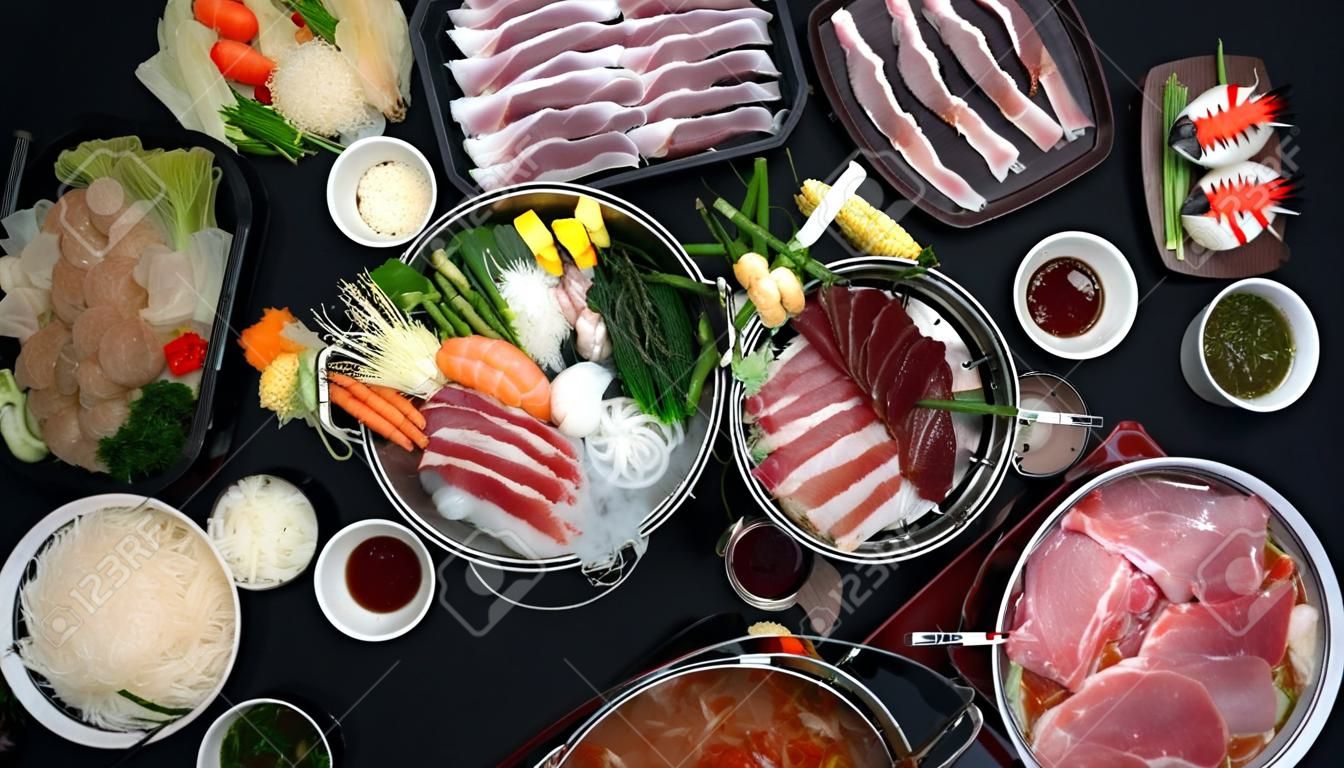 신선한 얇게 썬 고기, 해산물, 검은 배경을 가진 야채, 일본 요리와 함께 냄비에 샤부샤부를 먹는 사람들의 상위 뷰