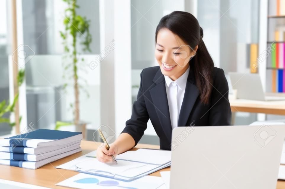 Donna d'affari asiatica millenaria allegra e attraente o consulente finanziario femminile che si concentra sui suoi compiti, lavorando al rapporto alla sua scrivania.