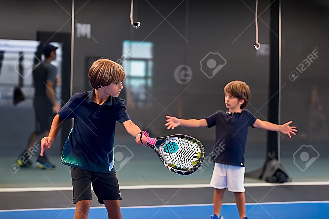 자녀에게 패들 수업을 가르치는 모니터, 그의 학생 - 트레이너는 어린 소년에게 실내 테니스 코트에서 패들 치는 방법을 가르칩니다.