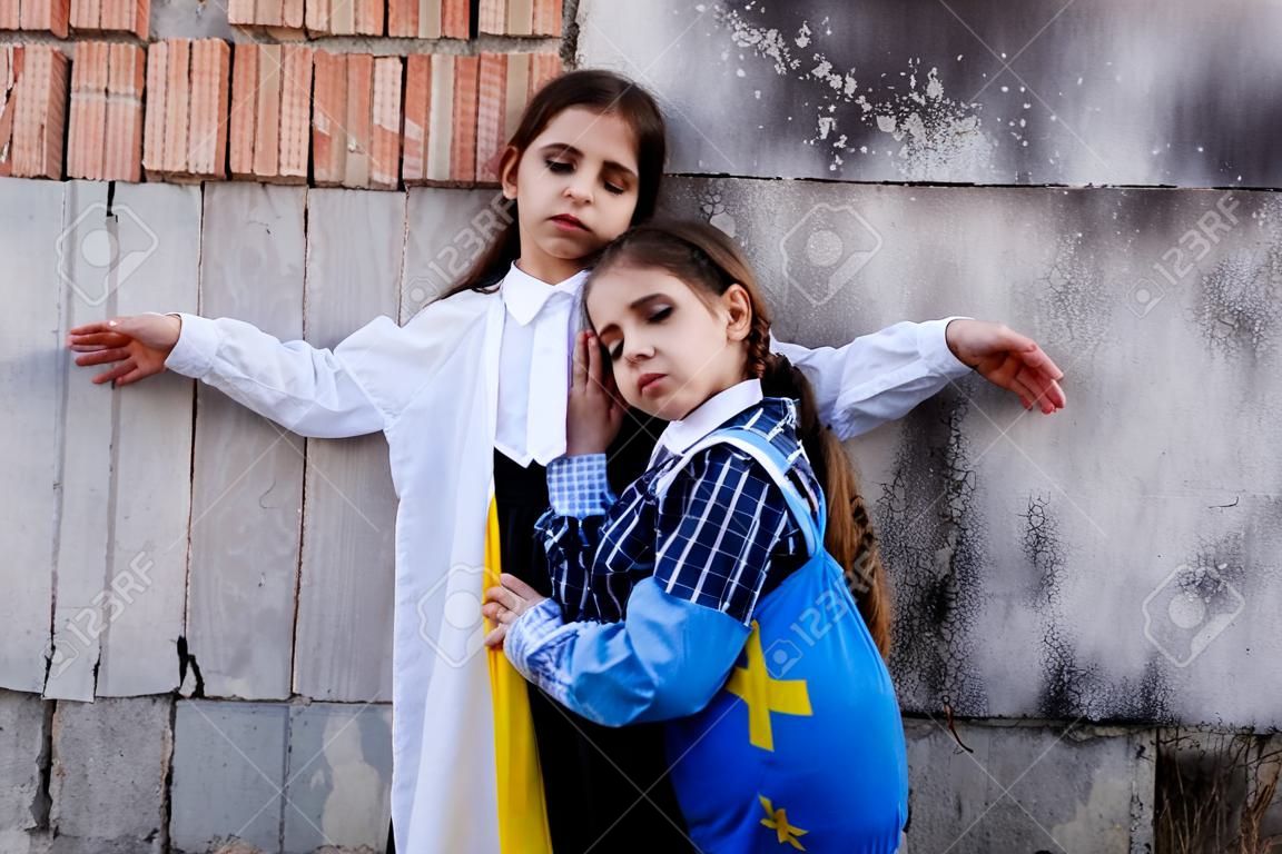 Meninas pequenas com bandeira ucraniana na frente de uma parede destruída a partir de bombas. As meninas agitam a bandeira nacional enquanto rezam pela paz