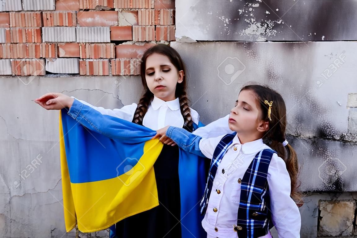 Meninas pequenas com bandeira ucraniana na frente de uma parede destruída a partir de bombas. As meninas agitam a bandeira nacional enquanto rezam pela paz