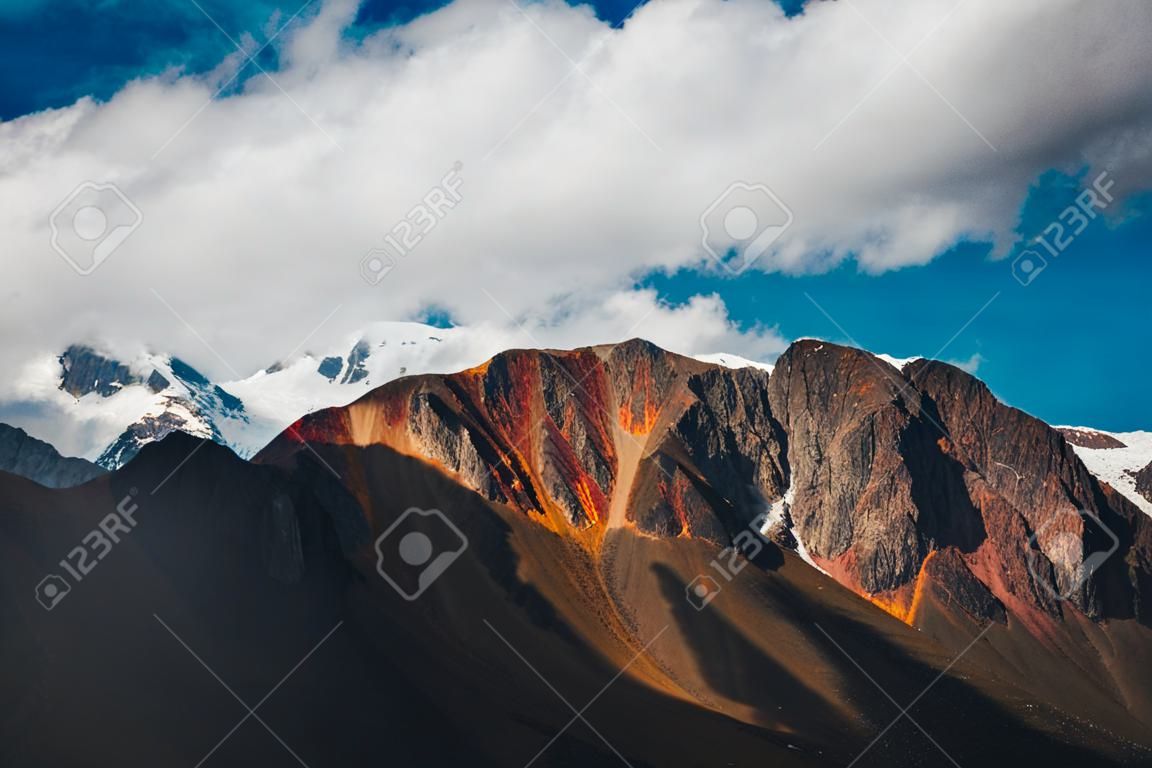 햇빛 아래 화려한 갈색 붉은 오렌지색 산벽 뒤에 눈 덮인 산봉우리가 있는 아름다운 고원 풍경. 높은 생생한 갈색 붉은 오렌지색 산과 큰 눈 꼭대기가 있는 맑은 산의 풍경