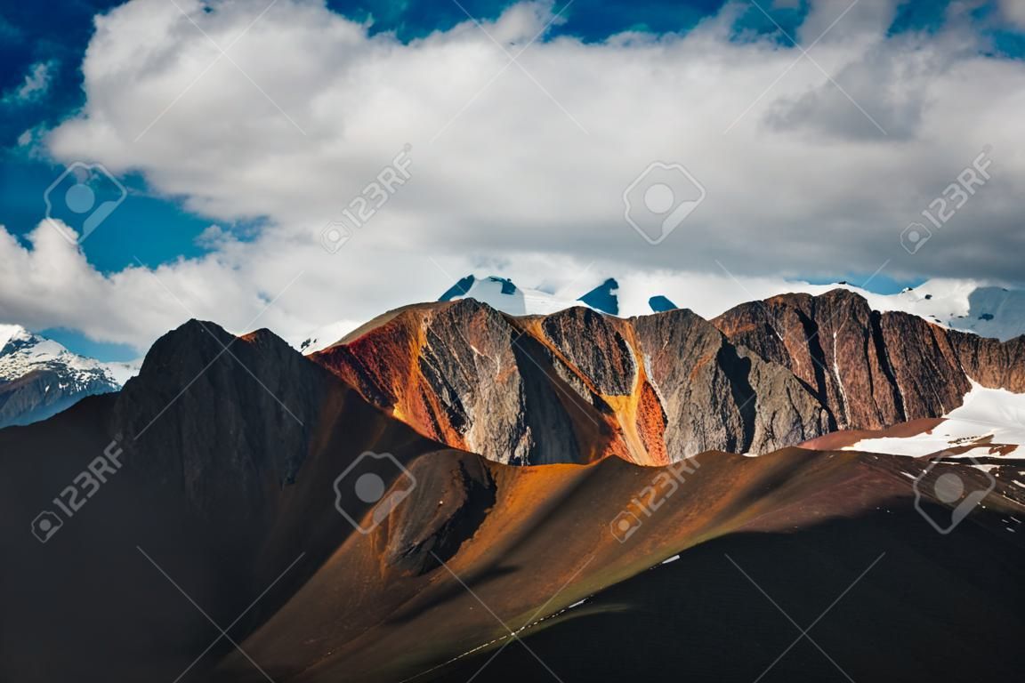 Scenic highlands landscape with great snowy mountain peak behind colorful brown red orange mountain wall in sunlight. Sunny mountains scenery with high vivid brown red orange mountain and big snow top