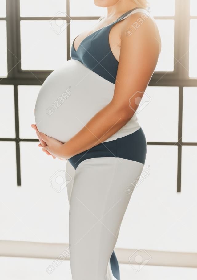 Un bel corpo di donna incinta, con la donna che tiene e sostiene delicatamente lo stomaco incinta. Dolce maternità femminile. Fotografato da uno sfondo bianco della finestra di luce naturale.