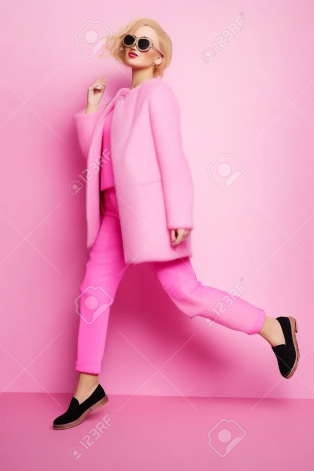foto de estudio de moda de mujer joven y hermosa con el pelo rizado rubio que viste elegante abrigo de color rosa, una blusa y gafas de sol luxuus