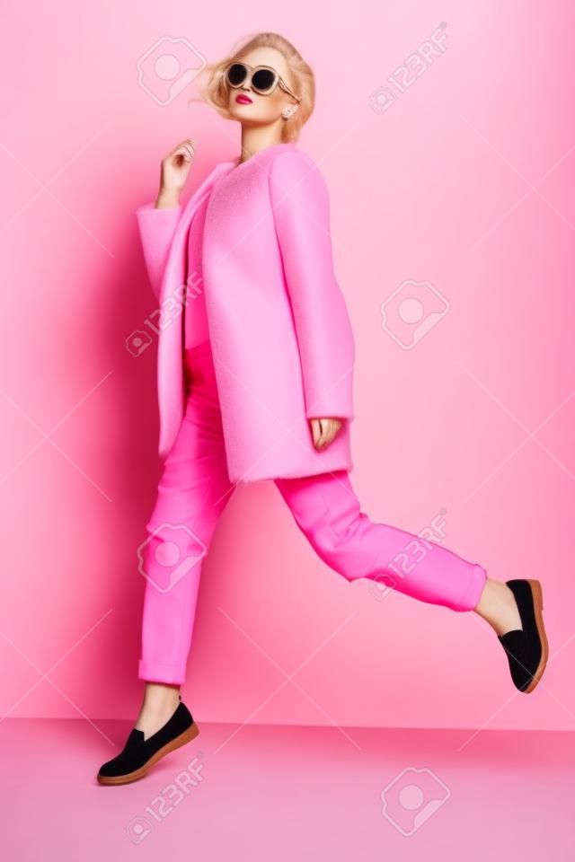 foto de estudio de moda de mujer joven y hermosa con el pelo rizado rubio que viste elegante abrigo de color rosa, una blusa y gafas de sol luxuus