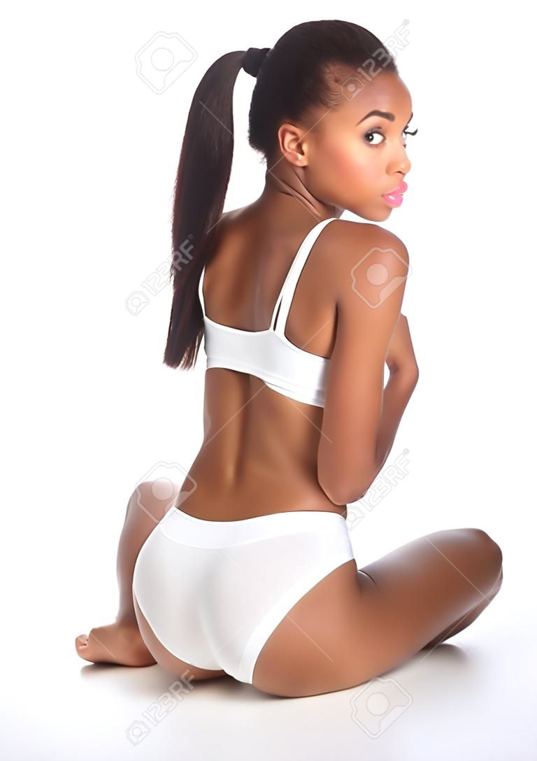 Vista posterior de una hermosa saludable joven mujer afroamericana vistiendo ropa deportiva blanca Mostrar cuerpo fit, sentado en el piso mirando hacia atrás sobre su hombro.