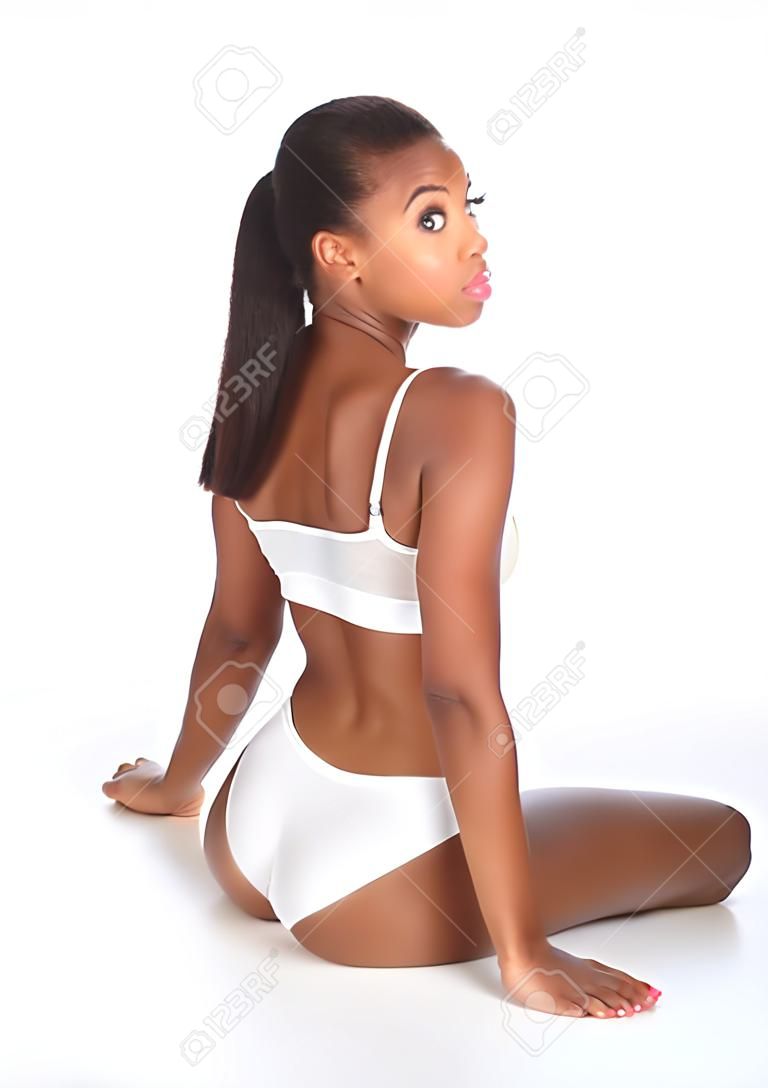 Vista posterior de una hermosa saludable joven mujer afroamericana vistiendo ropa deportiva blanca Mostrar cuerpo fit, sentado en el piso mirando hacia atrás sobre su hombro.