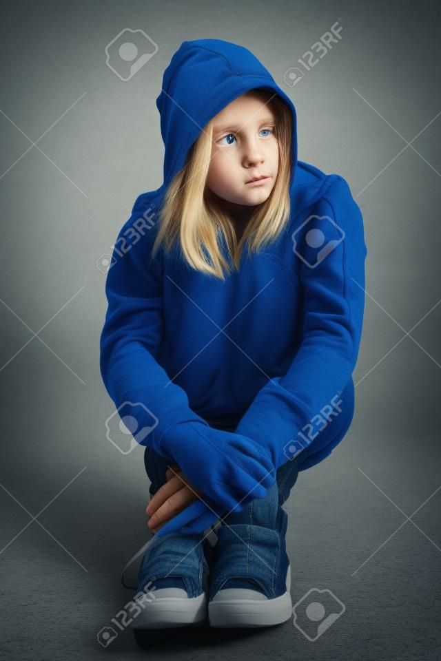 Hoodie on for distressed and frightened young blonde teenager girl sitting on floor looking scared and alone with big sad eyes, wearing jeans and blue jumper.