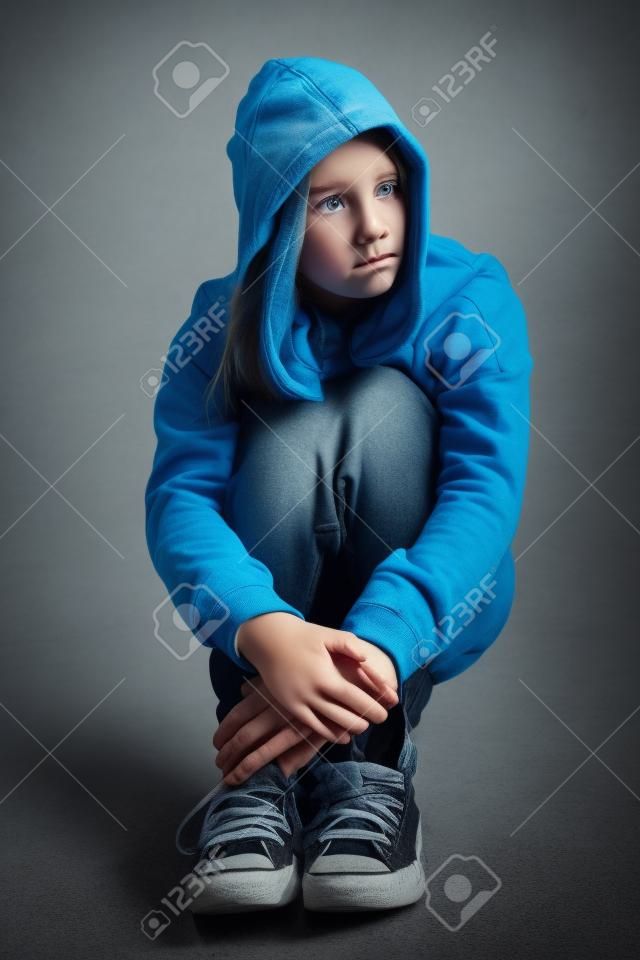 Felpa con cappuccio sopra per difficoltà e paura giovane ragazza adolescente bionda seduta sul pavimento cercando spaventata e sola, con grandi occhi tristi, indossa jeans e maglione blu.