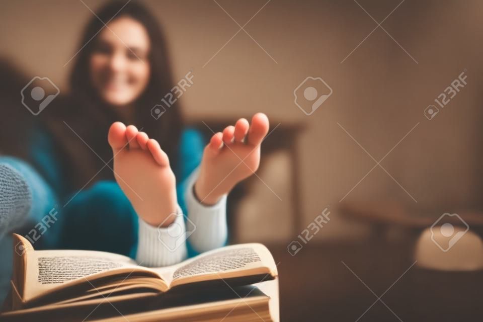 Joven mujer morena feliz con libro usando suéter en casa. Cerrar de pies