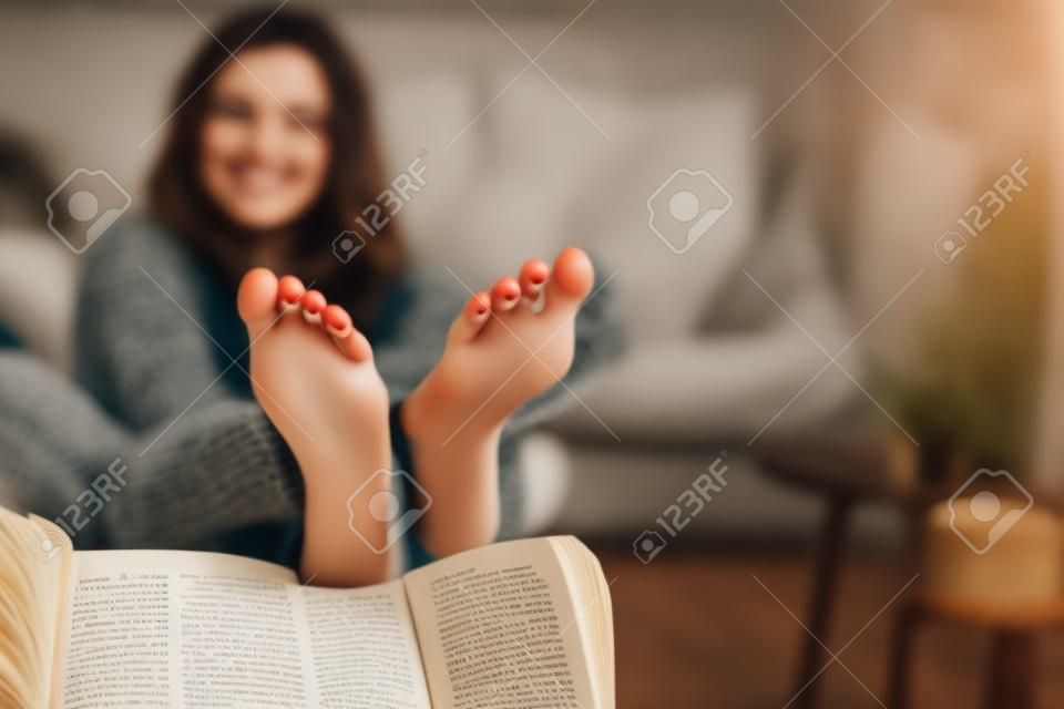 Joven mujer morena feliz con libro usando suéter en casa. Cerrar de pies