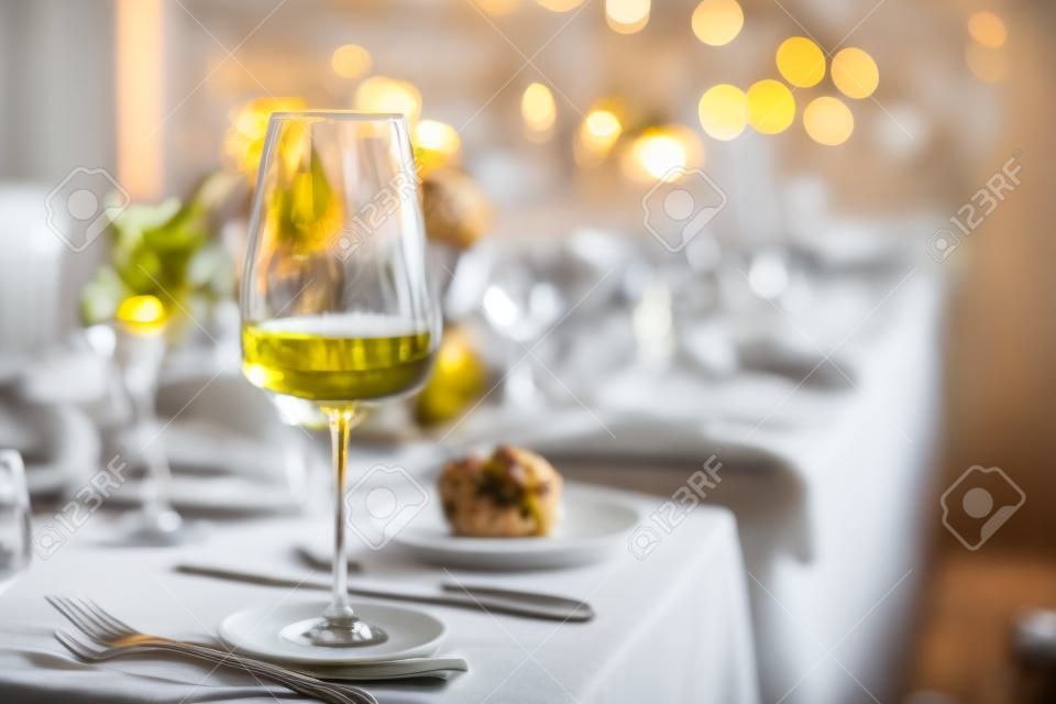 Selective focus on tall glass with white wine on foreground, standing against blurred background of uncleaned dinner table after festive banquette with remaining service settings, food and drinks