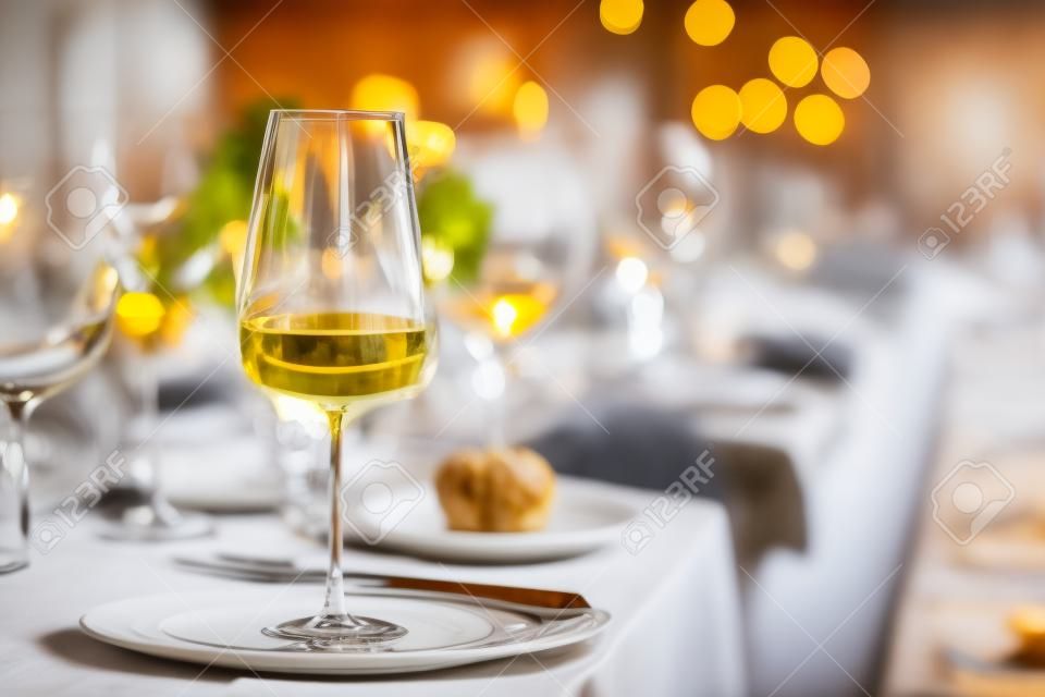 Selective focus on tall glass with white wine on foreground, standing against blurred background of uncleaned dinner table after festive banquette with remaining service settings, food and drinks