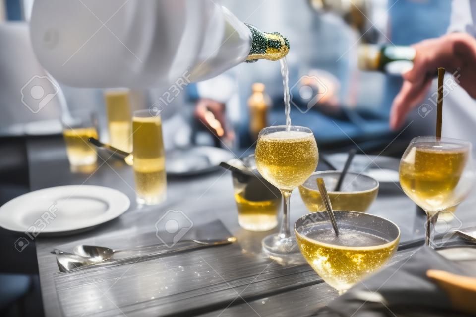 Ein Mann gießt ein Glas Champagner ein, Nahaufnahme fließender Sektgerichte, Snacks, Teller, Soßen, Menschen, die im Hintergrund speisen, ein freundliches Mittagessen oder eine Party in einem Café oder Restaurant