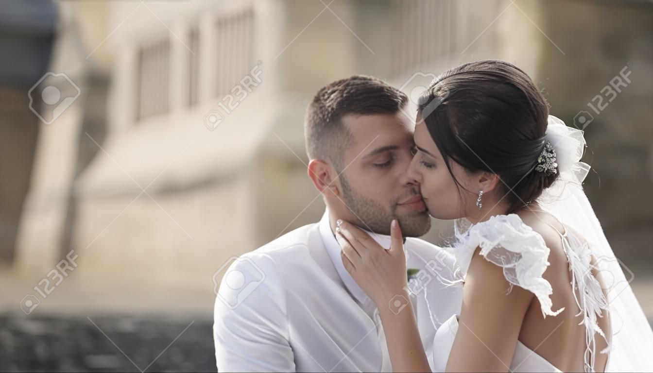 Close-up of beautiful fairytale bride in a fashionable white wedding dress and groom sitting in the bench, smiling and kissing. Action. A storybook wedding