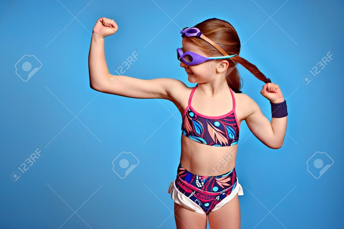 Menina forte gasta levanta o braço e olhando para os músculos, pronto para nadar, poses sobre fundo azul, usa óculos de proteção e maiô