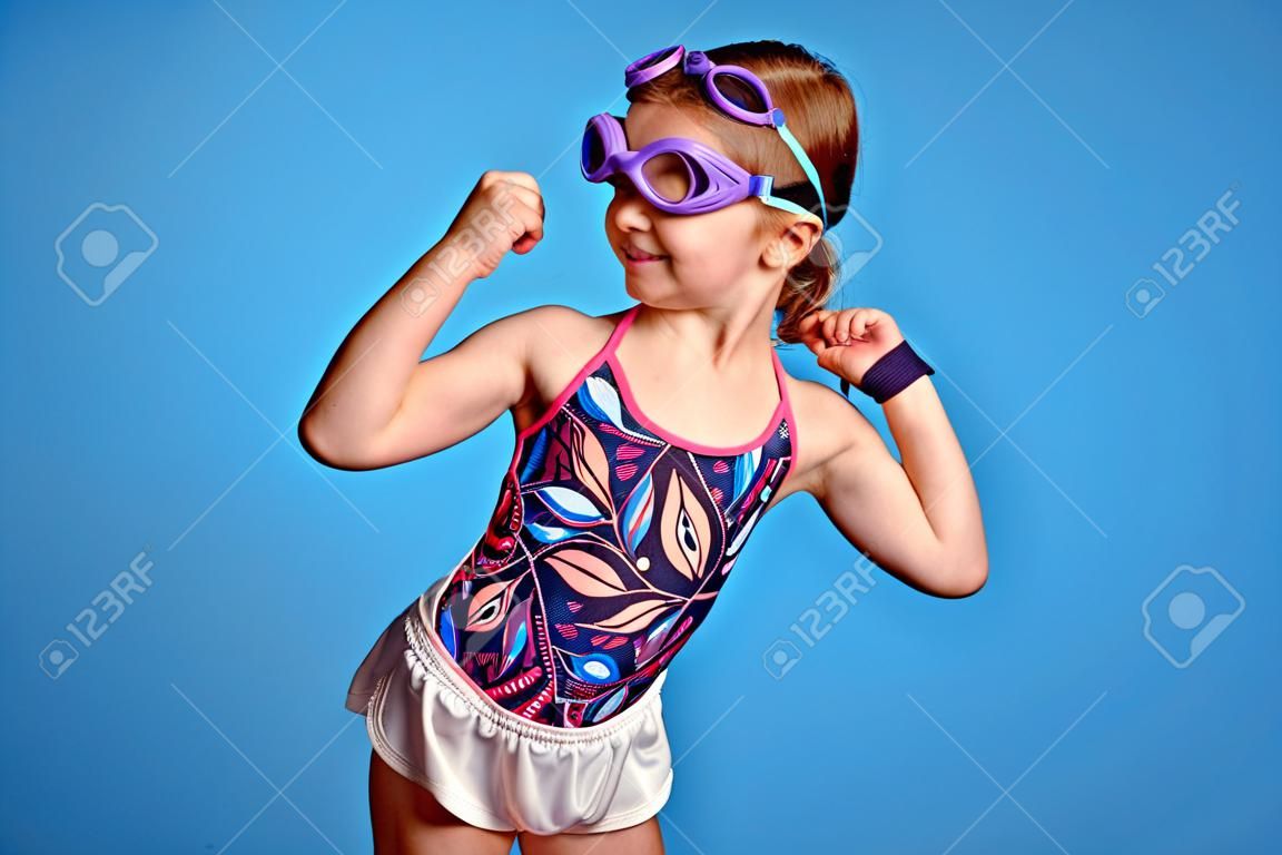 Menina forte gasta levanta o braço e olhando para os músculos, pronto para nadar, poses sobre fundo azul, usa óculos de proteção e maiô