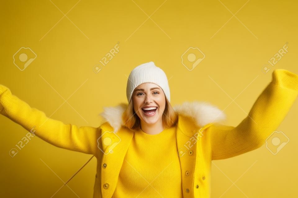 Retrato de mujer blanca sonriente, vistiendo una chaqueta y un gorro de punto, con fondo amarillo