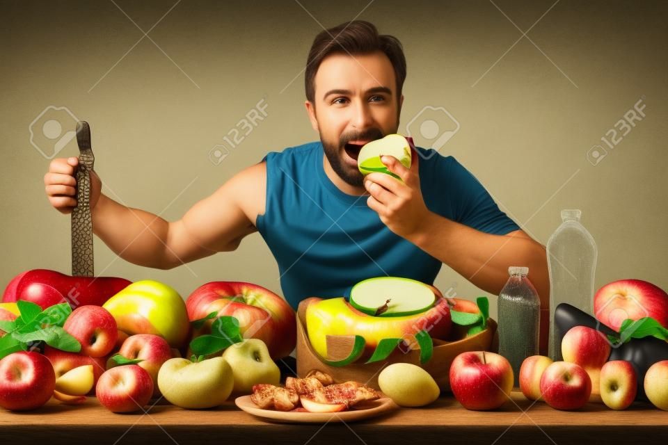 Sportsman eating apple showing fruits and vegetables, scales and accessories for sports on a table with venetian background.