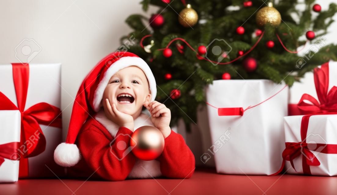 Wesołych Świąt i Nowego Roku. Portret dziecka w Santa czerwony kapelusz czeka na prezenty świąteczne. Mały chłopiec maluch leży pod choinką z prezentami i trzyma bombkę.