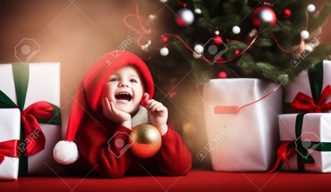 Happy xmas and New Year. Portrait of child in Santa red hat waiting for Christmas gifts. Little toddler boy lies under the Christmas tree with gifts and holds the Christmas ball.