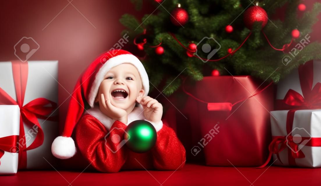 Happy xmas and New Year. Portrait of child in Santa red hat waiting for Christmas gifts. Little toddler boy lies under the Christmas tree with gifts and holds the Christmas ball.
