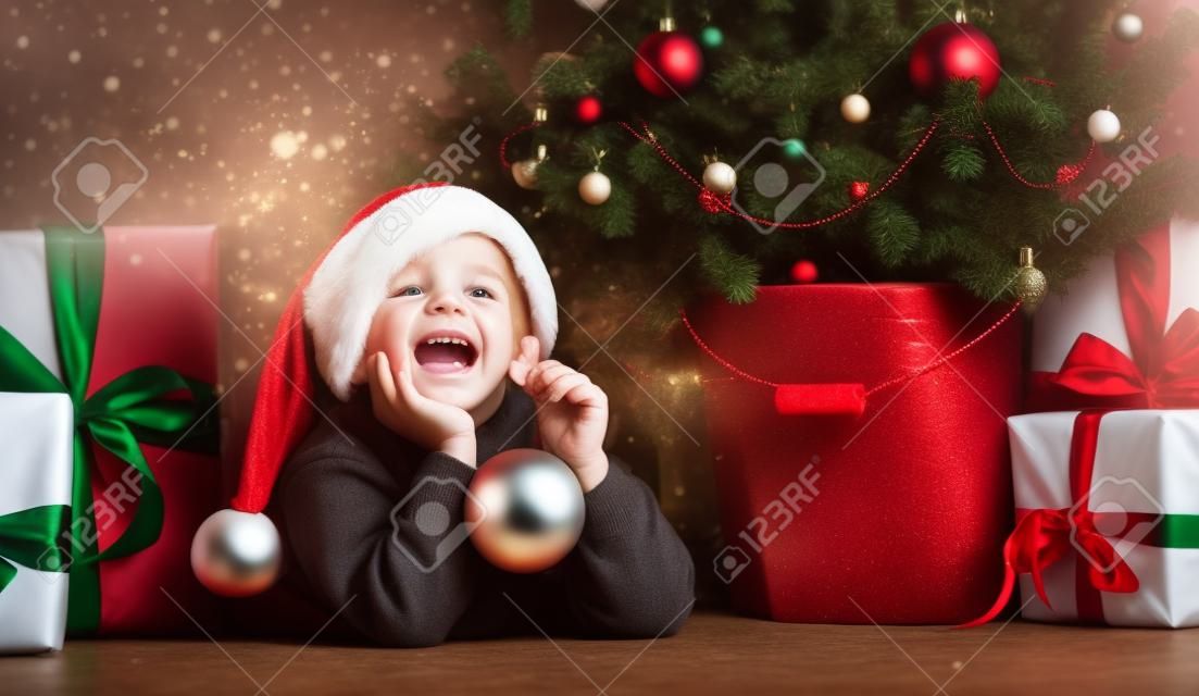 Wesołych Świąt i Nowego Roku. Portret dziecka w Santa czerwony kapelusz czeka na prezenty świąteczne. Mały chłopiec maluch leży pod choinką z prezentami i trzyma bombkę.