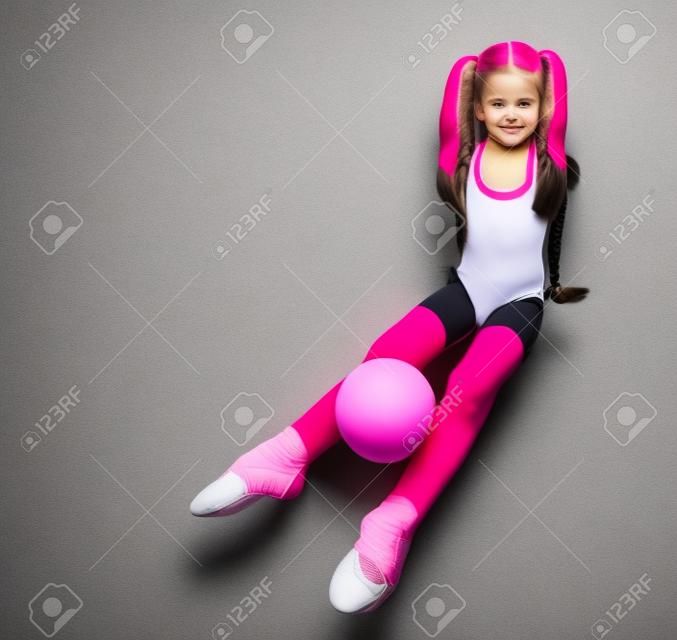 Cute girl gymnast with long hair in black sport body and uppers sitting on floor holding pink gymnastic ball between legs over white background, top view. Rhytmhic gimnastics beauty cocnept