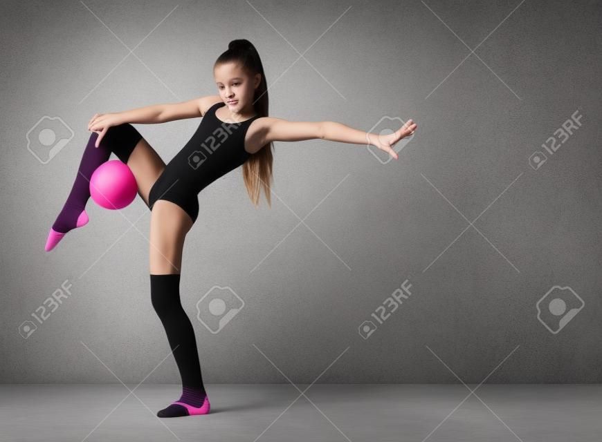 Młoda szczupła dziewczyna gimnastyczka w czarne legginsy ciało i skarpetki stojąc na jednej nodze i trzyma różową piłkę gimnastyczną z drugą nogą. Ćwiczenia gimnastyczne.