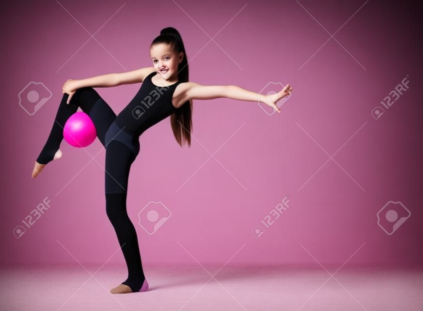 Młoda szczupła dziewczyna gimnastyczka w czarne legginsy ciało i skarpetki stojąc na jednej nodze i trzyma różową piłkę gimnastyczną z drugą nogą. Ćwiczenia gimnastyczne.