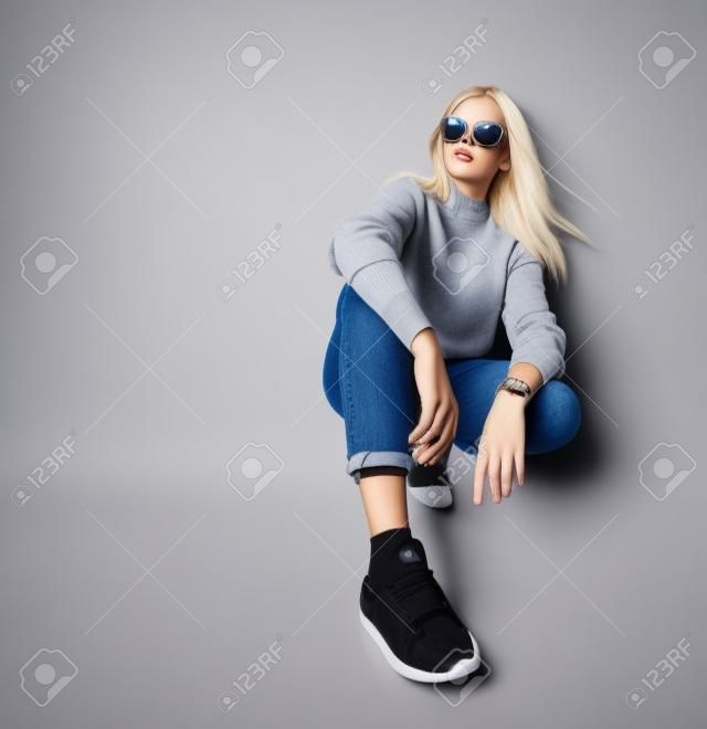 Blonde adolescent in sunglasses, smart watch, jeans, sweater, socks and sneakers. She squatting on floor, isolated on white studio background. Beauty, fashion, advertising. Copy space. Close up