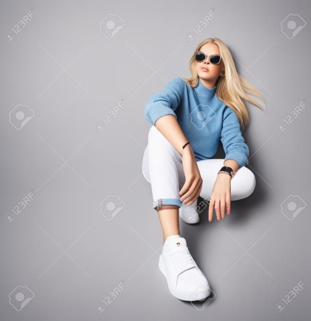 Adolescente blonde en lunettes de soleil, montre intelligente, jeans, pull, chaussettes et baskets. Elle accroupie sur le sol, isolée sur fond de studio blanc. Beauté, mode, publicité. Copiez l'espace. Fermer