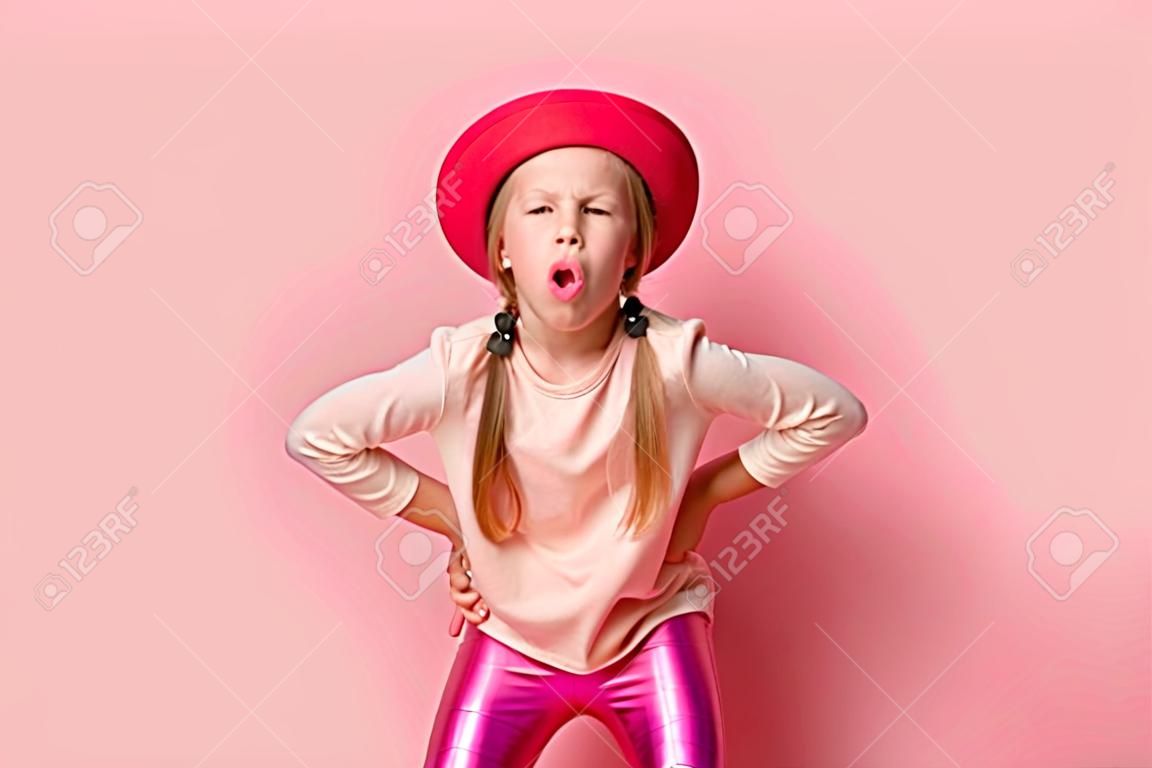 Mischievous little girl leaning forward with hands on hips and lolling while teasing or trolling someone. Close up shot isolated on pink. Stylish kids, childhood, emotions and gestures, games