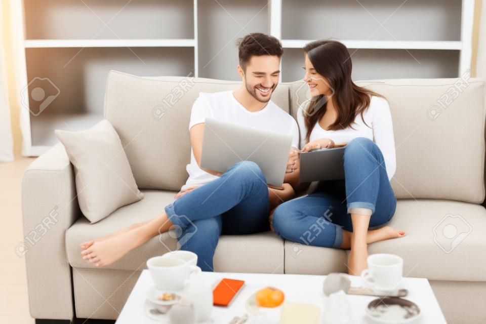 Cheerful happy couple is doing online shopping in internet at home indoors. They are on cozy beige couch in casual clothes, relaxing and buying goods easily