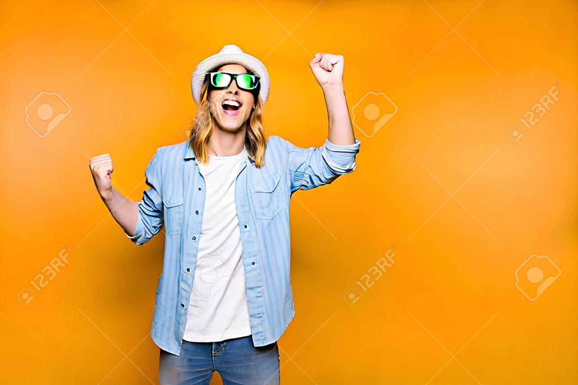 Ja! Der glückliche junge Hippie-Mann, der über gelbem Hintergrund, den tragenden Gläsern und dem Hut des glücklichen Mannes lokalisiert wird, wie er etwas mit erhobenen Händen, Gefühl, Erfolg, Geste und Leutekonzept gewann