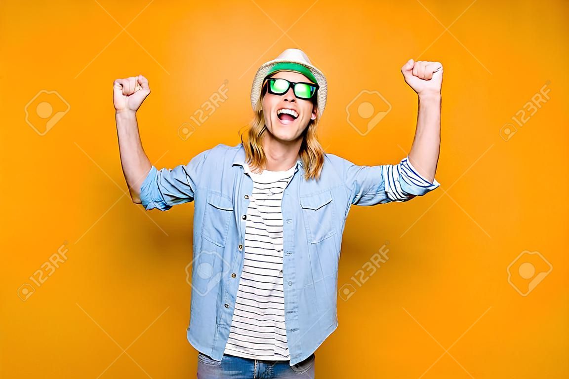 Ja! Der glückliche junge Hippie-Mann, der über gelbem Hintergrund, den tragenden Gläsern und dem Hut des glücklichen Mannes lokalisiert wird, wie er etwas mit erhobenen Händen, Gefühl, Erfolg, Geste und Leutekonzept gewann