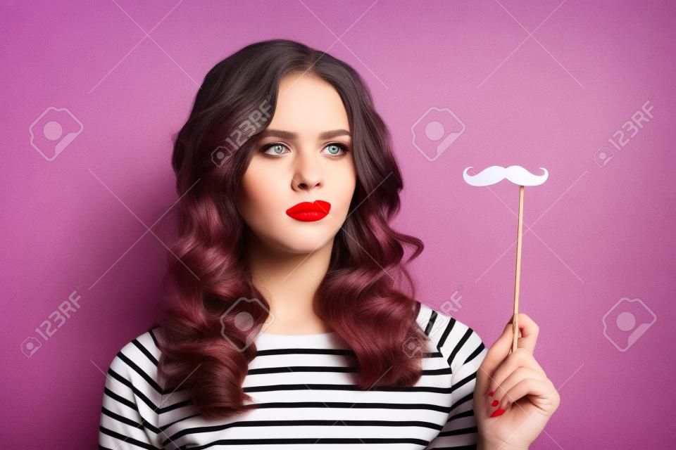 Portrait d'une fille confuse malheureuse avec du rouge à lèvres rouge regardant la moustache en papier découpé en carton noir sur bâton à la main isolé sur fond violet