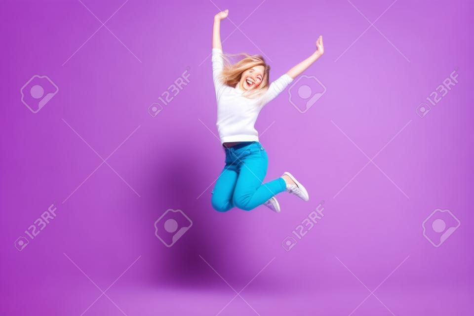Retrato da menina positiva alegre que salta no ar com punhos levantados que olham a câmera isolada no fundo violeta. Conceito da energia das pessoas da vida
