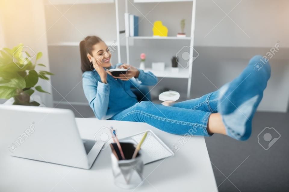 Bájos elegáns lány lábát az asztalra téve szünet szabadidejét használva okos telefon használata beszélgetés barátjával élvezi a pihenést szórakozás a munkahelyi munkaállomáson